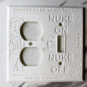 May include: A white rectangular light switch and outlet wall plate featuring embossed designs including a character giving a thumbs up, a mushroom cloud, and a rocket labeled USA. Text reads PROPERTY OF VAULT-TEC, NUKE ON, NUKE OFF, and FLIP RESPONSIBLY.