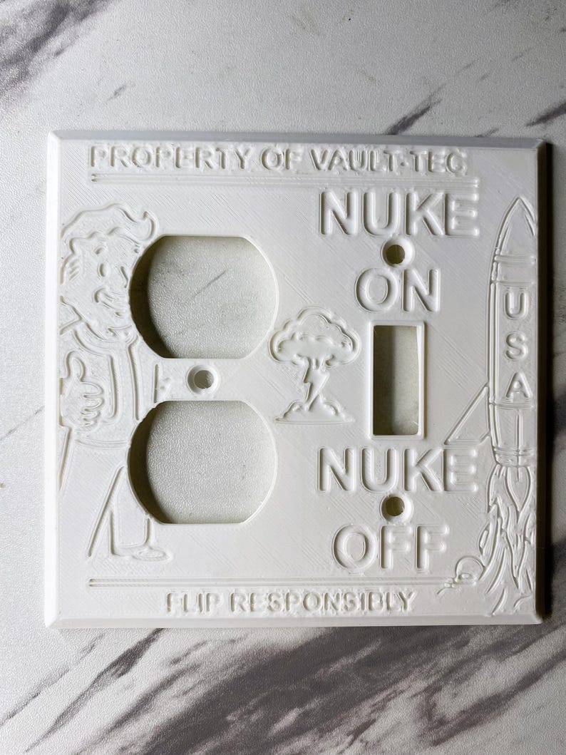 May include: A white rectangular light switch and outlet wall plate featuring embossed designs including a character giving a thumbs up, a mushroom cloud, and a rocket labeled USA. Text reads PROPERTY OF VAULT-TEC, NUKE ON, NUKE OFF, and FLIP RESPONSIBLY.
