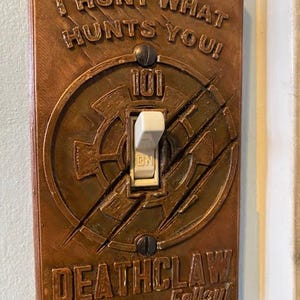 May include: A bronze-colored light switch cover with the text "I HUNT WHAT HUNTS YOU!" and "DEATHCLAW Fallout." The cover features a circular design with a white toggle switch in the center, marked "ON."