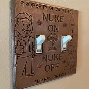 May include: A copper-colored double light switch plate featuring the text PROPERTY OF VAULT-TEC, NUKE ON, NUKE OFF, and FLIP RESPONSIBLY. The plate displays an embossed Vault Boy character, a mushroom cloud, and a rocket labeled USA.