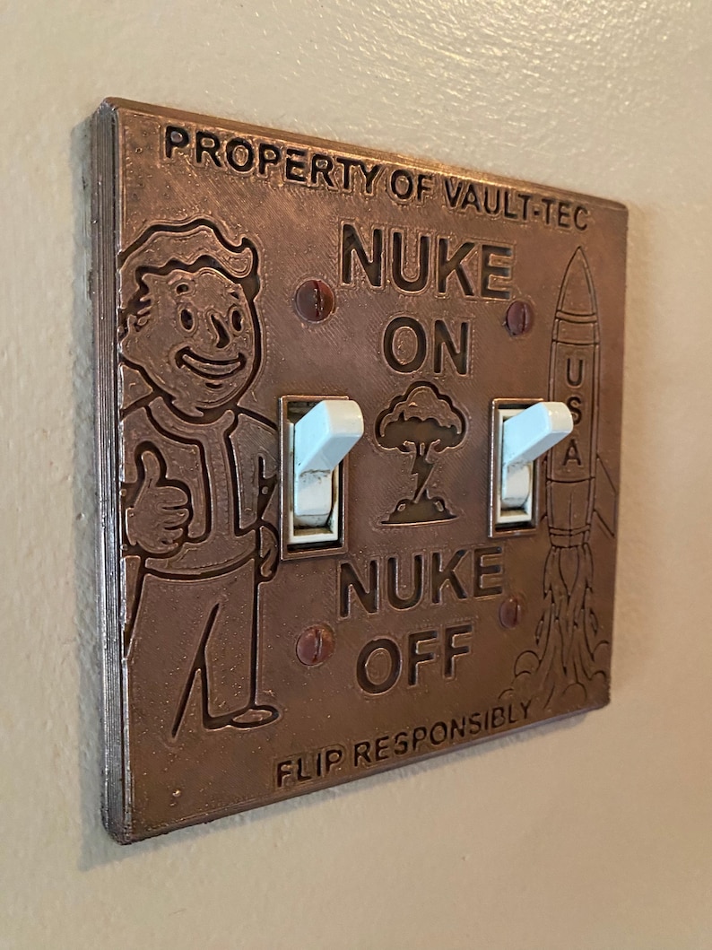 May include: A copper-colored double light switch plate featuring the text PROPERTY OF VAULT-TEC, NUKE ON, NUKE OFF, and FLIP RESPONSIBLY. The plate displays an embossed Vault Boy character, a mushroom cloud, and a rocket labeled USA.