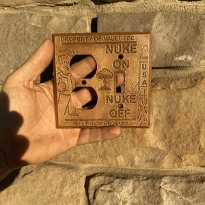 May include: A copper-colored rectangular light switch wall plate featuring embossed designs including a cartoon character, a rocket, and a mushroom cloud. The text reads PROPERTY OF VAULT-TEC, NUKE ON, NUKE OFF, USA, and FLIP RESPONSIBLY.