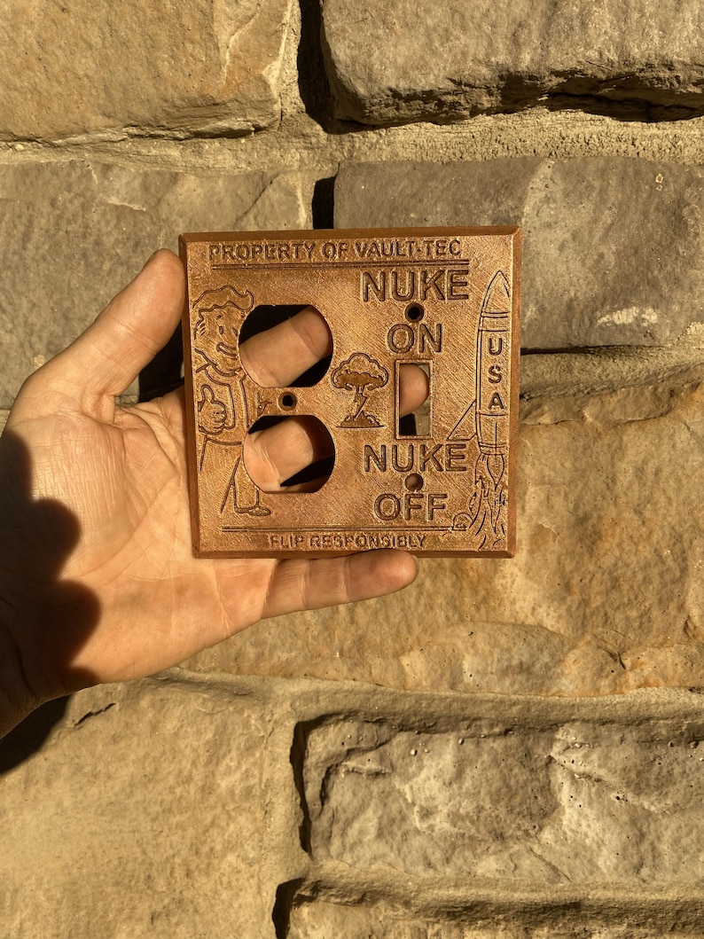 May include: A copper-colored rectangular light switch wall plate featuring embossed designs including a cartoon character, a rocket, and a mushroom cloud. The text reads PROPERTY OF VAULT-TEC, NUKE ON, NUKE OFF, USA, and FLIP RESPONSIBLY.