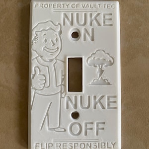 May include: A white rectangular light switch cover featuring embossed text and graphics. The top reads PROPERTY OF VAULT-TEC, the center shows NUKE ON and NUKE OFF with a cartoon character giving a thumbs up and a mushroom cloud, and the bottom reads FLIP RESPONSIBLY.