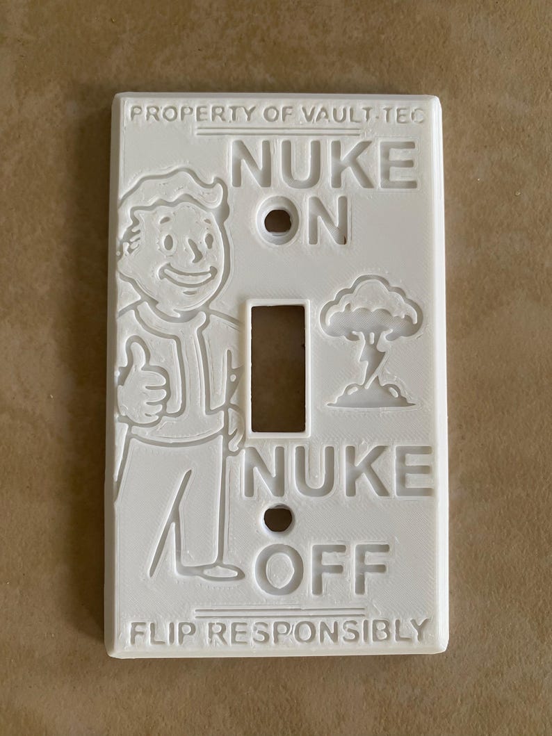 May include: A white rectangular light switch cover featuring embossed text and graphics. The top reads PROPERTY OF VAULT-TEC, the center shows NUKE ON and NUKE OFF with a cartoon character giving a thumbs up and a mushroom cloud, and the bottom reads FLIP RESPONSIBLY.