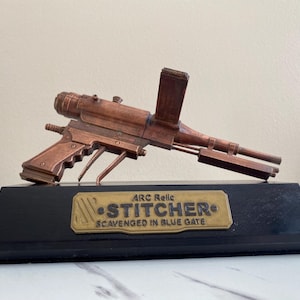 May include: A copper-colored, futuristic-looking "Stitcher" weapon model, mounted on a black base. The model has intricate details and is labeled "ARC Relic" and "Scavenged in Blue Gate" on a brass-colored plaque.