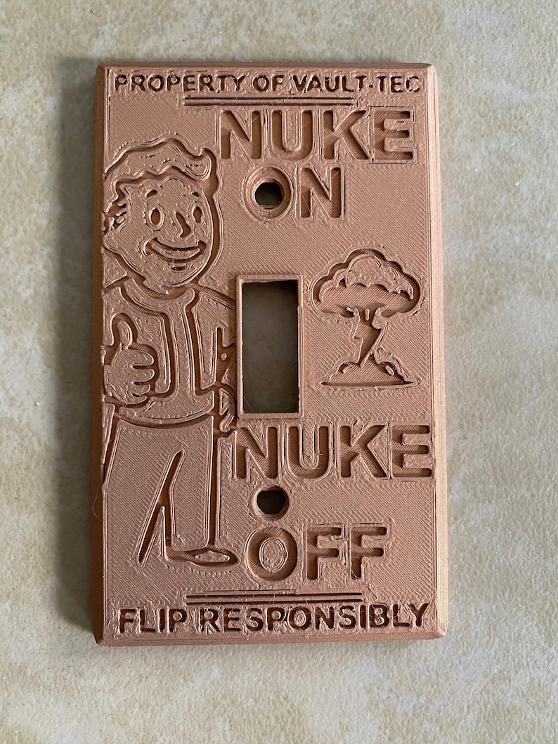 May include: A light brown rectangular light switch cover featuring embossed text and graphics. The top reads PROPERTY OF VAULT-TEC, the center features a character giving a thumbs up and a mushroom cloud, with NUKE ON and NUKE OFF text, and the bottom reads FLIP RESPONSIBLY.