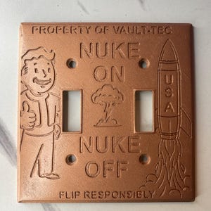 May include: A copper-colored double light switch plate featuring embossed text reading PROPERTY OF VAULT-TEC, NUKE ON, NUKE OFF, and FLIP RESPONSIBLY. The design includes a cartoon character giving a thumbs up, a mushroom cloud, and a rocket labeled USA.
