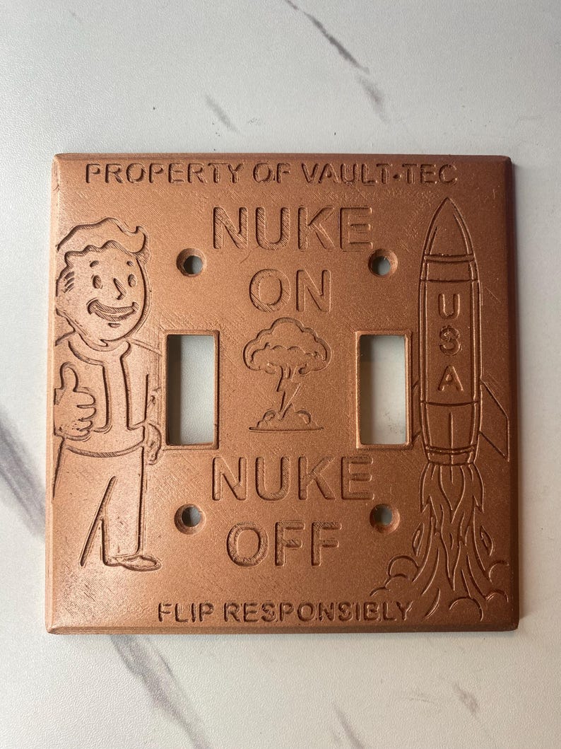 May include: A copper-colored double light switch plate featuring embossed text reading PROPERTY OF VAULT-TEC, NUKE ON, NUKE OFF, and FLIP RESPONSIBLY. The design includes a cartoon character giving a thumbs up, a mushroom cloud, and a rocket labeled USA.