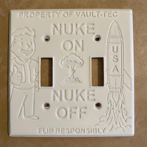 May include: A white rectangular double light switch cover featuring embossed designs including a cartoon character giving a thumbs up, a rocket labeled USA, a mushroom cloud, and the text PROPERTY OF VAULT-TEC, NUKE ON, NUKE OFF, and FLIP RESPONSIBLY.