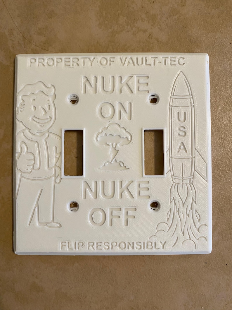 May include: A white rectangular double light switch cover featuring embossed designs including a cartoon character giving a thumbs up, a rocket labeled USA, a mushroom cloud, and the text PROPERTY OF VAULT-TEC, NUKE ON, NUKE OFF, and FLIP RESPONSIBLY.