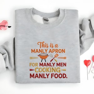 May include: A light gray sweatshirt with the embroidered text "This is a MANLY APRON FOR MANLY MEN - COOKING - MANLY FOOD." The design includes a grill, spatula, and other cooking utensils in orange and yellow.