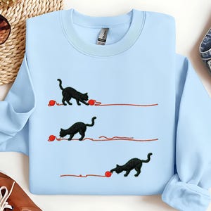 Puede incluir: Sudadera azul claro con gatos negros bordados jugando con ovillos de lana rojos. El diseño presenta tres gatos, cada uno con un ovillo y un hilo de lana. Cuello redondo, mangas largas.