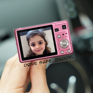 May include: A pink digital camera with a photo of a young girl on the screen. The camera has buttons and a lens. The text "DRIVE SAFE, DADDY" is printed below the screen.