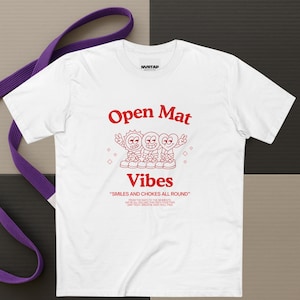 Puede incluir: Camiseta blanca con texto y gráficos rojos. La camiseta presenta las palabras "Open Mat Vibes" sobre una ilustración de dibujos animados de tres personajes sonrientes. Debajo, la frase "SMILES AND CHOKES ALL ROUND". Un cinturón morado está en el fondo.