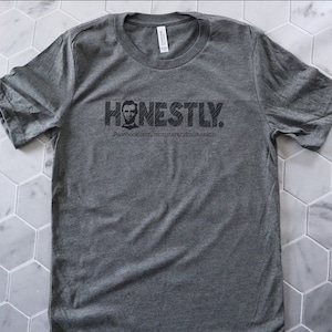 Op de afbeelding: Een heidegrijs T-shirt met een zwarte afbeelding van een mannengezicht en het woord "HONESTLY." De tekst "facebook.com/honestlyandmore.com" is onder de afbeelding gedrukt.