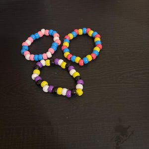 Könnte beinhalten: Drei Perlenarmbänder sind abgebildet. Ein Armband hat rosa, weiße und blaue Perlen. Ein anderes hat gelbe, rote, blaue und weiße Perlen. Das dritte Armband hat lila, schwarze, gelbe und weiße Perlen.