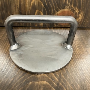 May include: A round, silver-colored metal burger press with a handle. The handle is a rectangular shape, welded to the circular base. The metal has a brushed finish. The press is designed for making burgers.