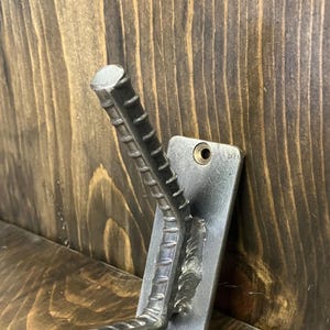 Puede incluir: Un gancho de pared de metal hecho de varilla corrugada, soldado a una placa rectangular. El gancho está en ángulo y tiene un extremo cilíndrico. La placa tiene dos agujeros para el montaje. El fondo es una superficie de madera.