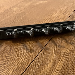 May include: A black metal wall-mounted coat rack with four rebar hooks. The hooks are attached to a rectangular metal plate. The rack is designed for hanging coats, hats, or other items. The rack is suitable for a hallway or entryway.