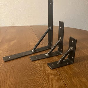 May include: Three industrial-style metal shelf brackets in varying sizes. Each bracket features a flat base and vertical support, connected by a diagonal rebar-style brace. The brackets are a dark gray color and have pre-drilled holes for mounting.