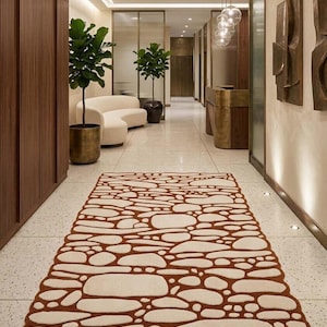 May include: A rectangular rug with a pebble design in cream and brown tones. The rug is placed in a hallway with a modern interior design, featuring plants, a sofa, and decorative wall art.