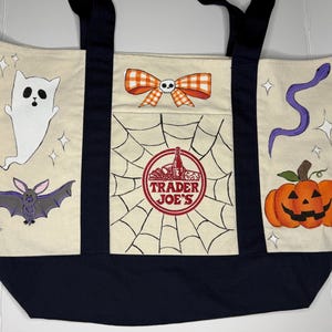 Może przedstawiać: Kremowa torba typu tote z granatowymi akcentami i uchwytami, z ilustracjami o tematyce Halloween. Zawiera ducha, nietoperza, dynię i pajęczynę z logo Trader Joe's. Świąteczny i praktyczny dodatek.