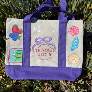 以下が含まれることがあります： 紫色のハンドルとトリムが付いたキャンバスのトートバッグ。 イチゴ、ブルーベリー、花、貝殻、巻貝、ヒトデのイラストが描かれています。 Trader Joe'sのロゴがバッグの中央に配置されています。