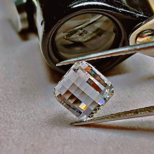 May include: A square-cut, faceted gemstone held by tweezers, reflecting light. The clear gem has multiple facets, displaying various colours. A jeweller's loupe is in the background, indicating a close inspection of the stone.