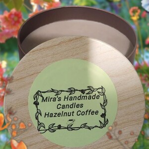 May include: A round candle in a brown container with a wooden lid. The lid has a green sticker with the text "Mira's Handmade Candles Hazelnut Coffee" in black. The background is a blurred image of flowers.