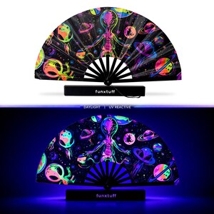 May include: A folding hand fan featuring a black background with a vibrant, multi-colored space theme including aliens, planets, and stars. The fan displays the text funxtuff on its base and is shown in both daylight and UV reactive lighting.