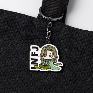 May include: A black metal keychain featuring a white-bordered enamel charm of a cartoon character with long brown hair, wearing a green cloak and reading a book. The text INFJ is printed in black on the left side next to a small yellow lantern.