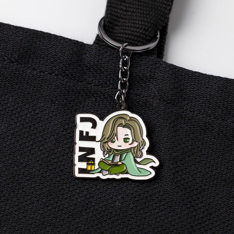 May include: A black metal keychain featuring a white-bordered enamel charm of a cartoon character with long brown hair, wearing a green cloak and reading a book. The text INFJ is printed in black on the left side next to a small yellow lantern.