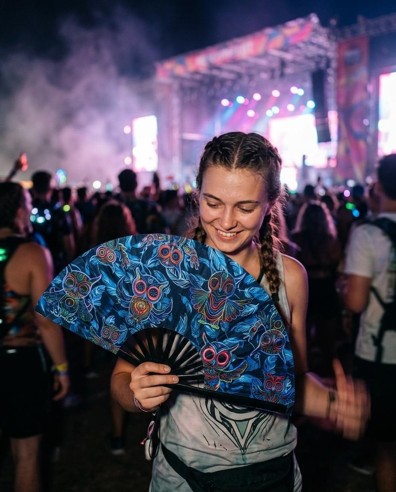 May include: A handheld folding fan featuring a dark blue fabric canopy decorated with a repeating pattern of stylized owls in vibrant neon colors including pink, orange, and green, attached to a black plastic frame with visible ribs.
