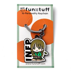 May include: A metal keychain featuring a chibi-style character with brown hair and a green cardigan, sitting and holding a book. The charm is attached to a silver ring on a white backing card with orange circles and the text funxtuff 16-Personality Keychain.