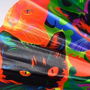 May include: A folding hand fan featuring a vibrant, psychedelic print of cat faces in bold shades of orange, black, blue, and green. The fabric surface displays a repeating pattern of feline portraits with stylized features and high-contrast colors.