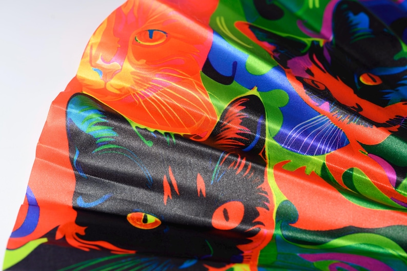 May include: A folding hand fan featuring a vibrant, psychedelic print of cat faces in bold shades of orange, black, blue, and green. The fabric surface displays a repeating pattern of feline portraits with stylized features and high-contrast colors.