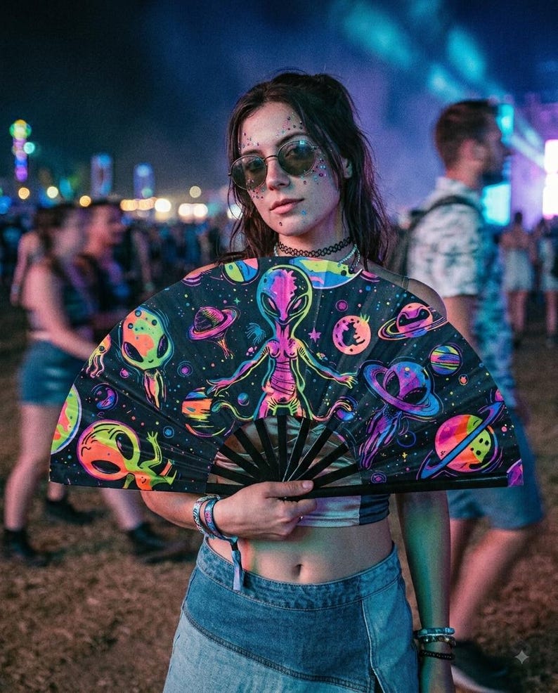 May include: A handheld folding fan featuring a black background with a vibrant, neon-colored graphic print of aliens, planets, and celestial bodies. The fan has black ribs and a wide, semi-circular shape when fully opened for display.