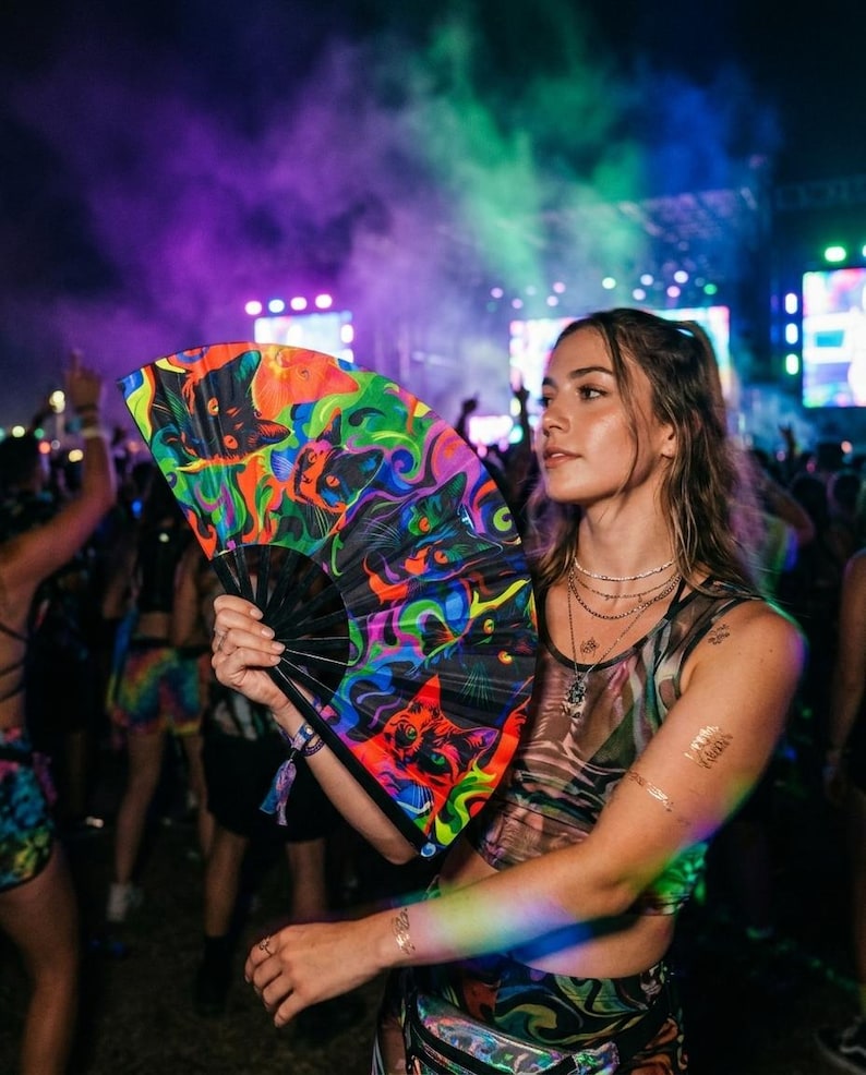 May include: A folding hand fan featuring a vibrant, psychedelic graphic print of cat faces in neon shades of green, orange, and blue against a black background, with a black frame and a wrist strap, held in an outdoor festival setting.