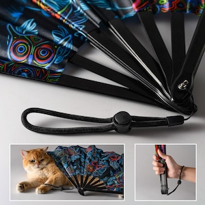May include: A folding hand fan with black plastic ribs and a fabric leaf featuring a vibrant, multicolored owl pattern on a black background. The fan includes a black wrist strap with an adjustable sliding toggle for secure carrying and handling.