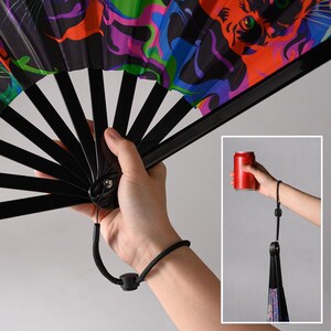 May include: A folding hand fan with black plastic ribs and a vibrant, multicolored graphic print featuring stylized cat faces in shades of orange, blue, purple, and green. The fan includes a black wrist strap with an adjustable plastic toggle.