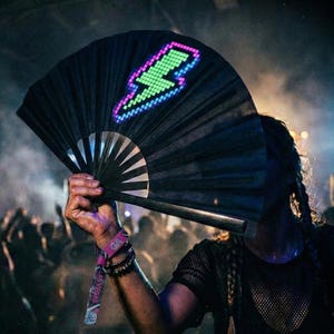 May include: A black folding hand fan featuring a pixelated lightning bolt graphic illuminated in bright green and purple. The fan has a dark frame and is held open, displaying the vibrant, glowing light pattern against the dark fabric surface.