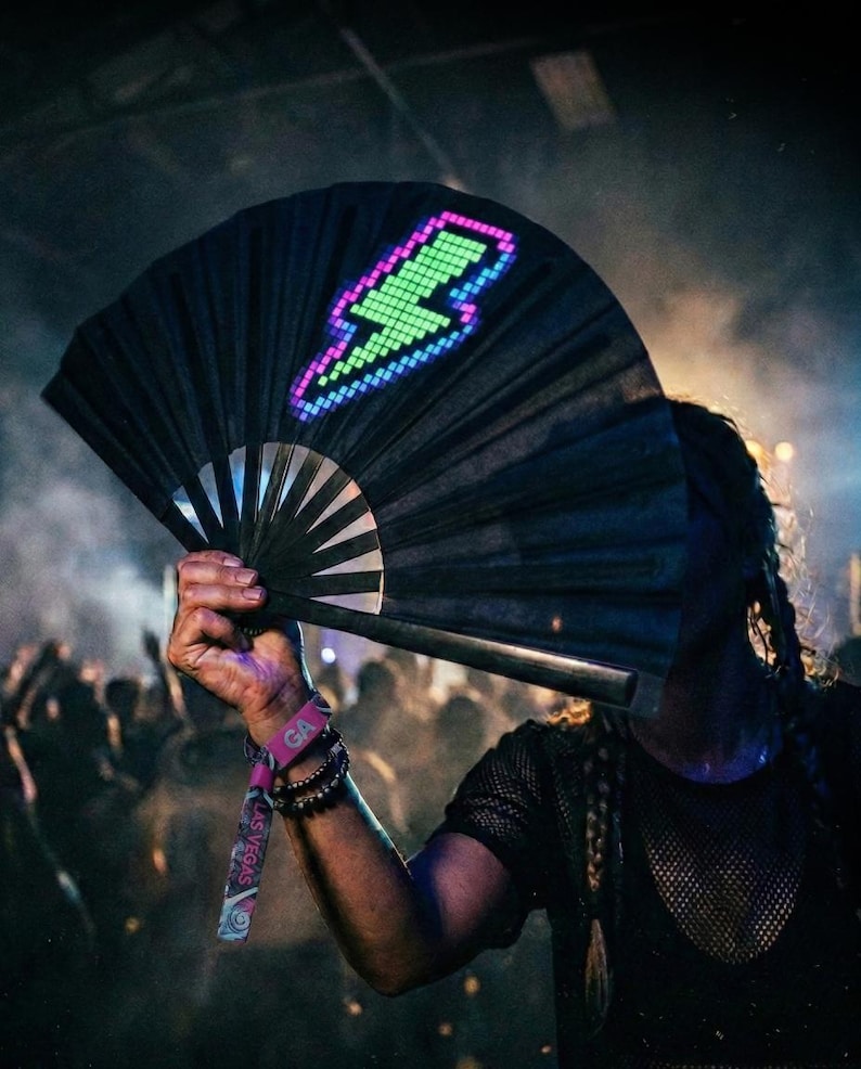 May include: A black folding hand fan featuring a pixelated lightning bolt graphic illuminated in bright green and purple. The fan has a dark frame and is held open, displaying the vibrant, glowing light pattern against the dark fabric surface.