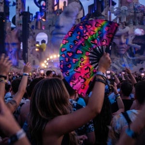 May include: A handheld folding fan featuring a vibrant rainbow gradient background decorated with a repeating pattern of black outlined hearts. The fan is constructed with black ribs and is held open in an upright position against a blurred background.