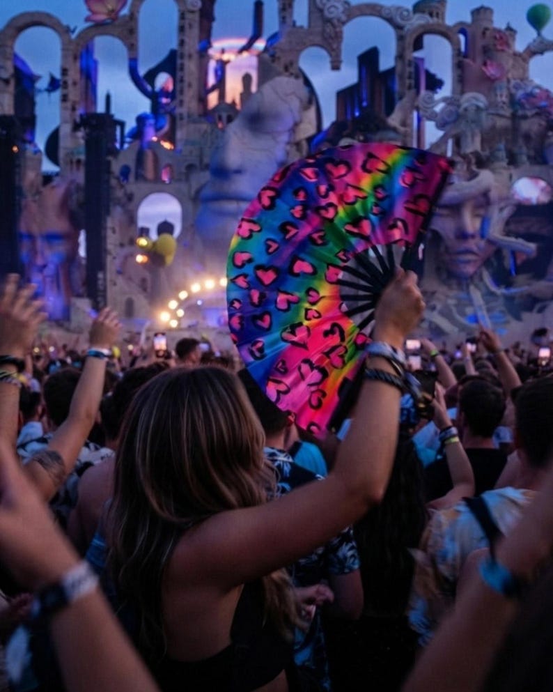May include: A handheld folding fan featuring a vibrant rainbow gradient background decorated with a repeating pattern of black outlined hearts. The fan is constructed with black ribs and is held open in an upright position against a blurred background.