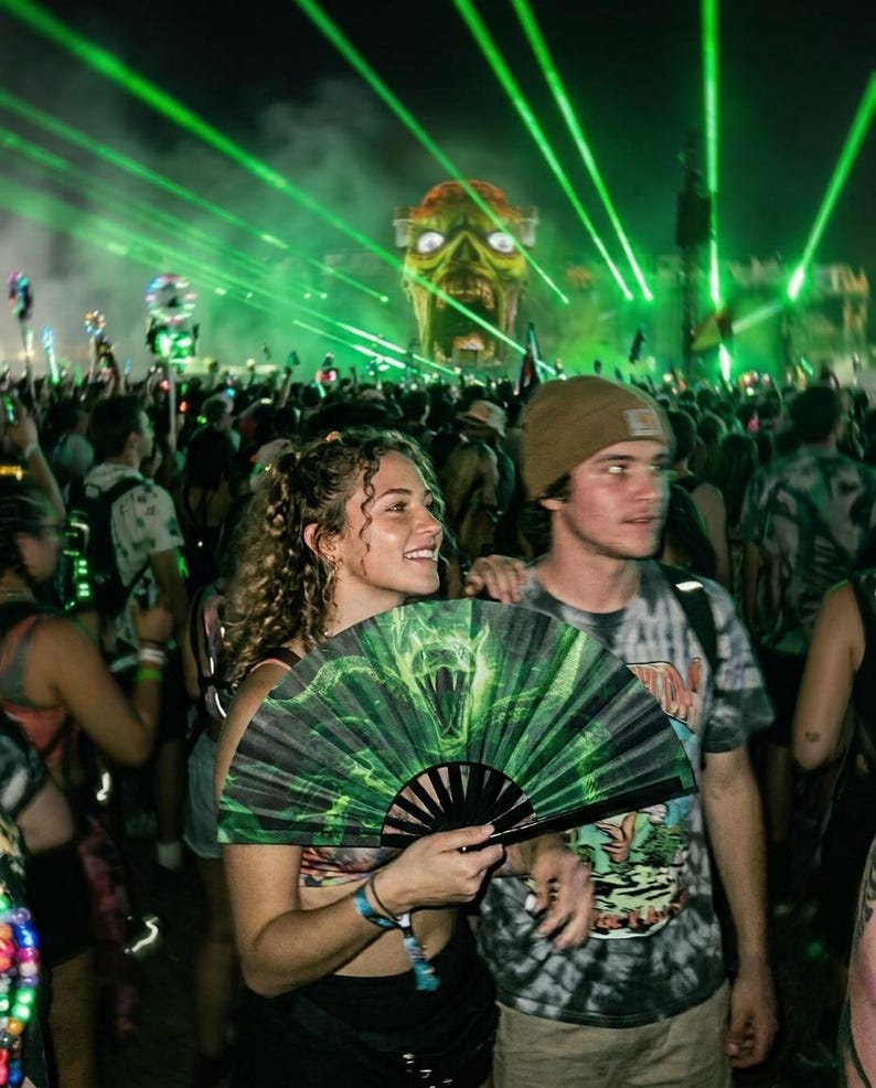 May include: A handheld folding fan featuring a green and black graphic print of a snake with bared fangs. The fan is held open, displaying the detailed illustration against a dark background with bright green laser lights and a large monster head.