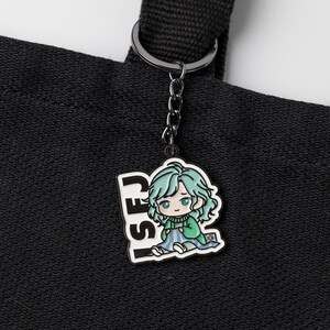 May include: A black metal keychain attached to a black fabric tote bag. The keychain features a colorful enamel charm depicting a cartoon character with light green hair, wearing a green sweater and blue blanket, with the text ISFJ in black.