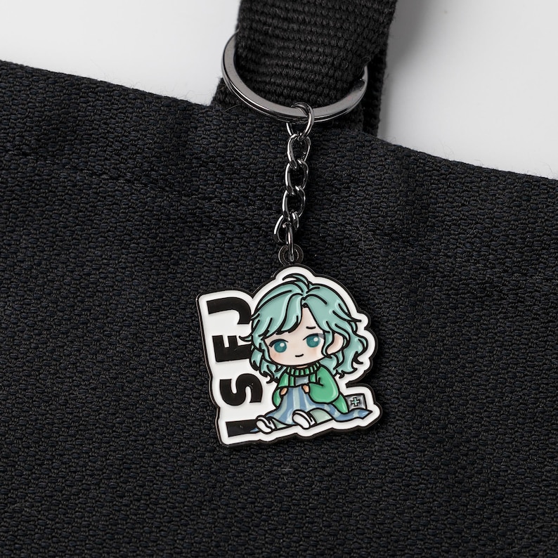 May include: A black metal keychain attached to a black fabric tote bag. The keychain features a colorful enamel charm depicting a cartoon character with light green hair, wearing a green sweater and blue blanket, with the text ISFJ in black.
