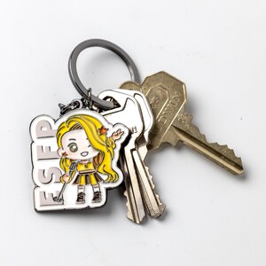 May include: A metal keychain featuring a white enamel charm with the text ESFP and a cartoon character with long yellow hair, a yellow top, and a brown skirt. The charm is attached to a dark metal ring holding two silver-toned and brass-toned keys.