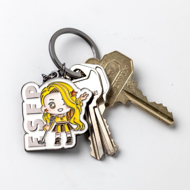 May include: A metal keychain featuring a white enamel charm with the text ESFP and a cartoon character with long yellow hair, a yellow top, and a brown skirt. The charm is attached to a dark metal ring holding two silver-toned and brass-toned keys.